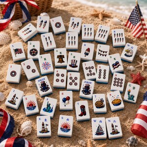 May include: A set of rectangular mahjong tiles featuring patriotic American-themed illustrations, including fireworks, flags, eagles, and landmarks, arranged on a sandy surface with seashells and small American flag decorations nearby.
