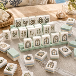 K&ouml;nnte beinhalten: Ein Set aus wei&szlig;en Mahjong-Spielsteinen mit gr&uuml;nen botanischen Illustrationen, blau-goldenen bogenf&ouml;rmigen Zahlendesigns und kreisf&ouml;rmigen Mustern. Einige Steine stehen auf hellgr&uuml;nen Kunststoffhaltern, andere liegen verstreut auf einer texturierten Oberfl&auml;che.