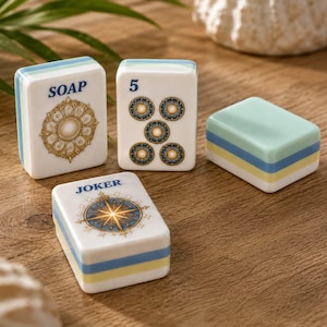 K&ouml;nnte beinhalten: Vier rechteckige Mahjong-Spielsteine mit wei&szlig;en Vorderseiten und geschichteten hellblauen, gelben und wei&szlig;en Seiten. Ein Stein zeigt das Wort SOAP und ein goldenes Blumenemblem, einer die Zahl 5 mit f&uuml;nf kreisf&ouml;rmigen Symbolen und einer JOKER mit einem Kompassstern.