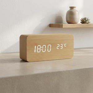 Puede incluir: Un reloj despertador digital rectangular con un acabado de grano de madera clara. La parte frontal cuenta con una pantalla LED blanca brillante que muestra la hora 18:00 y la temperatura de 23 grados Celsius. El reloj tiene esquinas redondeadas.