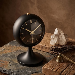 Peut inclure: Une horloge de table sphérique noir mat avec une base sur pied, dotée d'un cadran sombre, d'aiguilles dorées et d'index dorés. Le mot QUARTZ est imprimé en petites lettres blanches au centre inférieur du cadran de l'horloge.