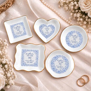 May include: Five white ceramic trinket dishes with gold-painted rims, featuring blue floral wreath designs. Each dish displays the text "sister EST. 1999" and "auntie EST. 2026" with a small bow icon in the center of the floral pattern.