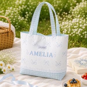 May include: A white tote bag with a light blue gingham pattern on the handles and base. The bag features a light blue bow print, a white lace trim, and the name AMELIA embroidered in blue. A small label reads A GOGO ORIGINAL.