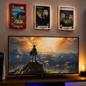Può includere: Un televisore a schermo piatto mostra un paesaggio di un videogioco con un personaggio su una roccia, posizionato sopra una console in legno. Tre copertine di videogiochi incorniciate sono montate sulla parete sopra la TV, con Zelda, Kingdom Hearts e Expedition 33.