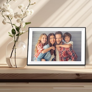 May include: A rectangular black picture frame containing a photograph of a family of four, including two children and two adults, positioned next to a clear glass vase filled with white flowers on a wooden surface.