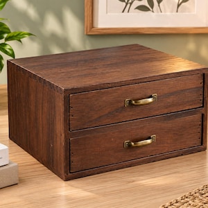 May include: A dark brown wooden desktop organiser featuring two horizontal drawers. Each drawer is fitted with a small, curved, antique-style brass metal handle. The unit has a rectangular shape with visible wood grain texture throughout.