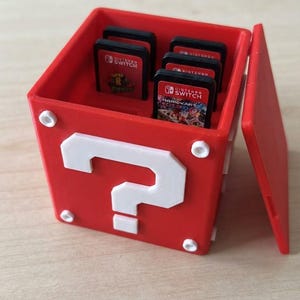 Peut inclure: Une boîte de rangement carrée rouge avec un point d'interrogation blanc sur le devant et les côtés, conçue pour contenir des cartouches de jeu Nintendo Switch. La boîte possède un couvercle amovible et contient plusieurs cartouches noires.