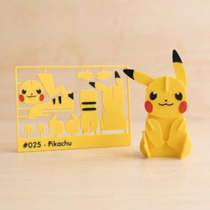 May include: A yellow plastic model kit featuring a Pikachu character, with a flat sprue frame labeled #025 - Pikachu containing various unassembled parts and a fully assembled yellow Pikachu figure with black-tipped ears and red cheeks.