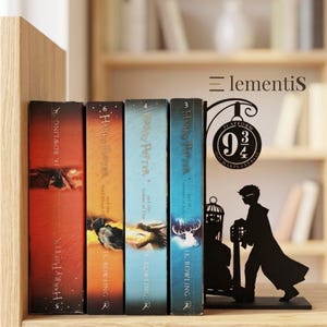 Puede incluir: Un sujetalibros de metal negro con la silueta de una persona empujando un carrito de equipaje hacia un letrero de Platform 9 3/4, junto a cuatro libros de Harry Potter. El sujetalibros incluye un diseño de lámpara y el texto Elementis.