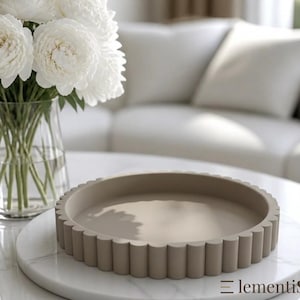 Könnte beinhalten: Ein rundes, beigefarbenes Dekotablett mit einem gewellten, geriffelten Randdesign. Das Tablett hat eine glatte, flache Innenfläche und eine gleichmäßige matte Oberfläche. Der Text "lementis" ist dunkel auf der unteren rechten Seite gedruckt.