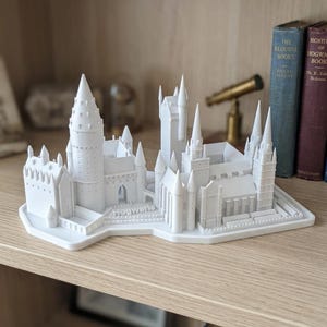 Puede incluir: Un modelo blanco impreso en 3D de un complejo de castillo que presenta múltiples torres, agujas y detalles arquitectónicos. La base de la estructura incluye la palabra Hogwarts en relieve sobre una sección plana de la plataforma del modelo.