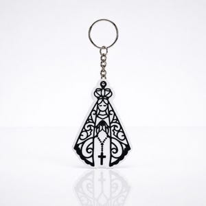 Peut inclure: Un porte-clés blanc avec une illustration au trait noir représentant une silhouette portant une couronne et une longue robe ornée, avec un chapelet suspendu aux mains. L'ensemble est fixé à un anneau en métal argenté par une chaînette.