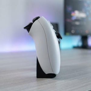 以下が含まれることがあります： 黒いプラスチック製のスタンドに立てかけられた白いビデオゲーム用コントローラー。コントローラーを側面から見た様子で、湾曲した白いシェル、黒いショルダーボタン、デバイスの上端付近に見える黒いアナログスティックが確認できます。