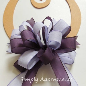 May include: A purple and lavender ribbon bow with tails, tied around a wooden circle. The bow is made of sheer ribbon with a subtle shimmer. The text "Simply Adornments" is visible below the bow.
