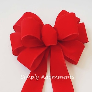 May include: A large, vibrant red velvet bow with long, flowing tails. The bow is centered and appears to be handmade, with a classic design. The text "Simply Adornments" is visible on the lower left side. This bow is suitable for holiday or gift decoration.