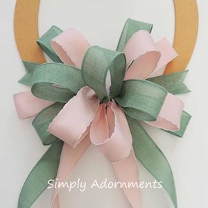 May include: A green and pink bow with a ruffled edge, tied on a wooden wreath form. The bow is made of two different types of fabric, one with a smooth texture and the other with a more textured look. The text "Simply Adornments" is visible at the bottom of the image.