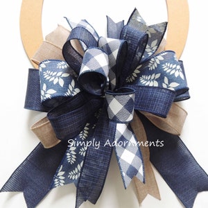 May include: A decorative bow featuring navy blue solid ribbon, navy blue ribbon with a white leaf pattern, and navy blue and white buffalo check ribbon. The bow is attached to a circular wooden frame and includes the text Simply Adornments.