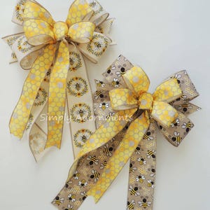 May include: Two decorative bows with bee and honeycomb patterns. One bow features a cream-coloured ribbon with yellow honeycomb and bee accents. The other bow has a tan ribbon with bee and honeycomb designs. The text "Simply Adornments" is visible.