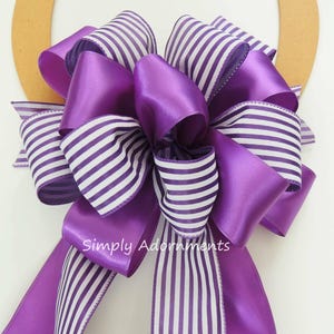 May include: Purple and white striped ribbon bow with a purple satin ribbon. The bow is tied with a loop and tails. The text "Simply Adornments" is printed on the bow.