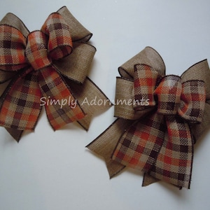 May include: Two decorative bows made of tan burlap fabric with a brown, orange, and cream plaid pattern on the center loops and tails. The edges of the ribbon are finished with dark brown stitching. The text Simply Adornments is visible.