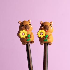 Può includere: Due bastoncini per capelli con figure di capibara marroni dall'espressione sorridente, ognuna delle quali tiene un fiore giallo con stelo verde. I capibara hanno piccole guance rosa e sono fissati su bastoncini di legno scuro, sottili e cilindrici.
