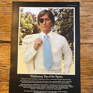 May include: A vintage advertisement for Hathaway shirts featuring a person wearing a white long-sleeved button-down shirt, a light blue necktie, and a black eye patch. The text reads Hathaway. Top of the Spots. Spot the Hathaway.