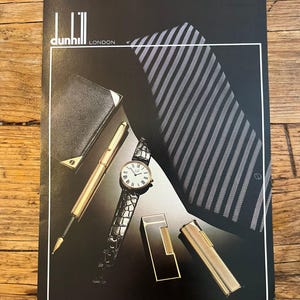 May include: A black advertisement poster for Dunhill London featuring a gold fountain pen, a wristwatch with a black textured strap, a black wallet, a striped tie, and two gold lighters. The text reads Dunhill London and Alfred Dunhill Ltd, 30 Duke Street, St. James's, London.
