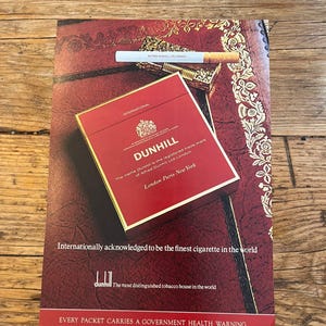 May include: A vintage advertisement for Dunhill cigarettes featuring a red pack with gold lettering, a single cigarette, and a gold lighter. The text reads Dunhill, International, London-Paris-New York, and Every packet carries a government health warning.