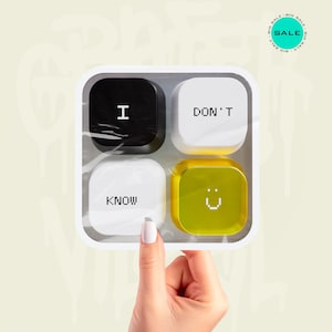 Op de afbeelding: Een vierkante plastic houder met vier afgeronde vierkante knoppen. De knop linksboven is zwart met de witte tekst I, rechtsboven wit met de tekst DON'T, linksonder wit met de tekst KNOW en rechtsonder geel met een smiley-gezichtje.