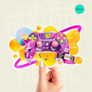 Puede incluir: Una pegatina que muestra un mando de videojuego con camuflaje morado y rosa, botones verde lima, un emblema de cara sonriente y el texto GAME OVER. El diseño incluye formas abstractas amarillas, un disco holográfico y una pulsera plateada PLAY GAME.