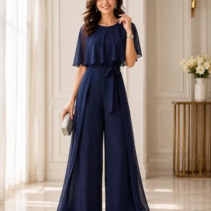 May include: A navy blue jumpsuit featuring a round neckline, short sheer cape-style sleeves, and wide-leg trousers. The garment includes a matching fabric tie belt at the waist and a layered chiffon overlay that drapes over the bodice and legs.