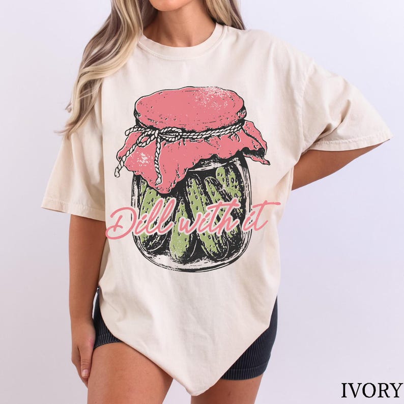 A woman wearing a white t-shirt with a graphic design of a jar of pickles and the text "Dell w/it" printed on it.