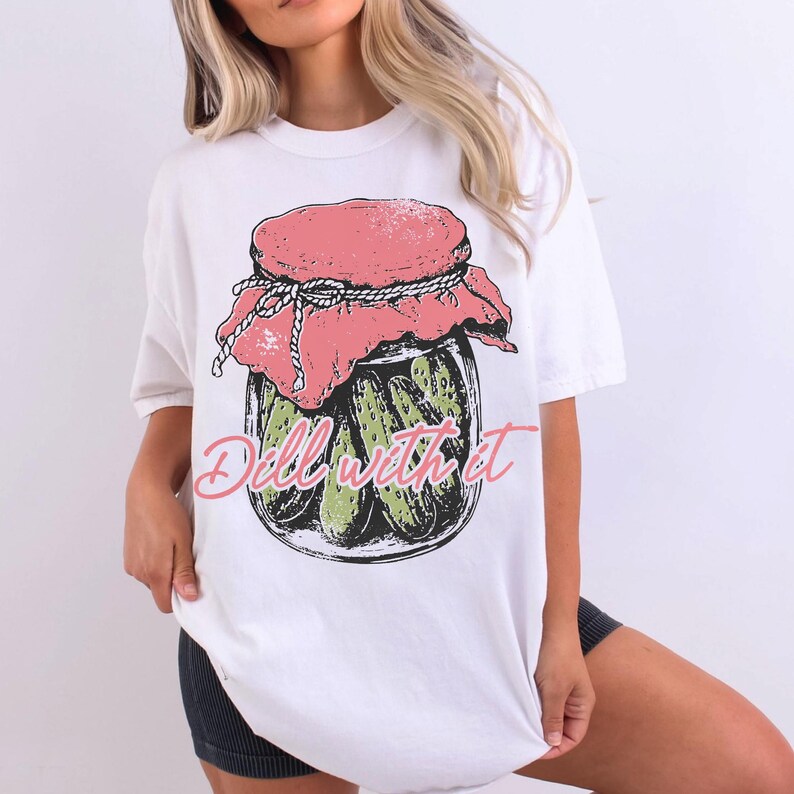 A woman wearing a white t-shirt with a graphic design of a jar of pickles and the text "Dill with it" printed on it.