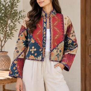 May include: A cropped, long-sleeved jacket featuring a patchwork design with various geometric and floral patterns in shades of red, navy blue, and beige. The jacket has a mandarin collar, an open front, and dark blue trim along the edges.