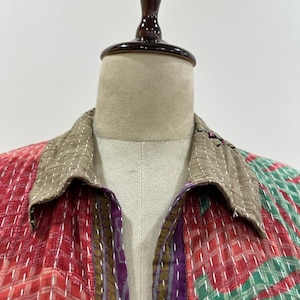 Può includere: Una camicia a maniche lunghe con colletto color marrone chiaro e un design patchwork con sezioni rosse, verdi e viola. Il tessuto è decorato con ricami a punto filza bianco su tutta la superficie, conferendo un aspetto strutturato.
