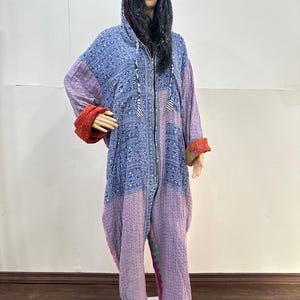 May include: A full-length hooded jumpsuit featuring a patchwork design with blue paisley patterns on the upper section and a light purple textured fabric on the lower section. The garment includes a front zipper, long sleeves with red cuffs, and a hood.