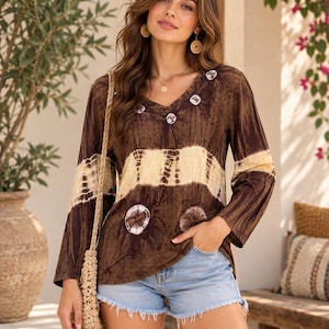 Puede incluir: Una blusa de manga larga con cuello en V y estampado tie-dye en tonos marrón y crema, con motivos circulares alrededor del cuello y la cintura, combinada con pantalones cortos de mezclilla azul claro con dobladillo deshilachado y bolso beige.