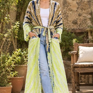 May include: A long, open-front duster cardigan featuring a tie-waist belt and two front pockets. The garment displays a tie-dye pattern in shades of olive green, bright lime green, and dark navy blue, worn over a white tank top and blue jeans.