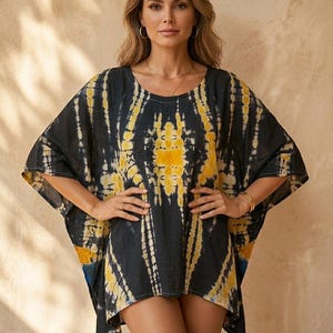 May include: A black and yellow tie-dye kaftan featuring a loose, flowing silhouette with wide sleeves and a round neckline. The garment displays a symmetrical pattern of vertical and abstract shapes in shades of mustard yellow and white against a dark black background.
