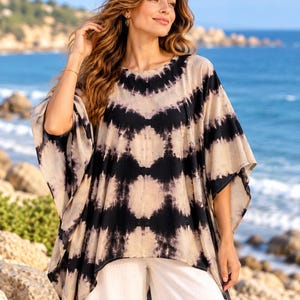 May include: A loose-fitting, short-sleeved poncho-style top featuring a black and beige tie-dye pattern with a repeating diamond motif. The garment has a round neckline and a wide, flowing silhouette that drapes over the shoulders and torso.