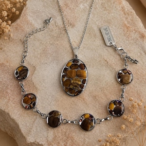 Könnte beinhalten: Ein silberfarbenes Halsketten- und Armbandset mit ovalen und runden Anhängern, die mit polierten braunen und goldenen Tigeraugen-Steinen gefüllt sind. Das Armband enthält ein kleines rechteckiges Metallschild mit der Gravur BELIEVE.