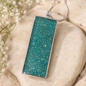 Peut inclure: Un collier pendentif rectangulaire avec une incrustation en résine bleu sarcelle remplie de fines particules de paillettes dorées. Le pendentif est enchâssé dans un cadre en métal argenté lisse et suspendu à une fine chaîne en métal argenté.