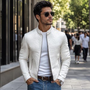 Puede incluir: Una chaqueta blanca con un patrón texturizado de piel de cocodrilo, que presenta un cuello alzado, una cremallera central plateada y dos bolsillos laterales. La chaqueta se lleva sobre una camiseta blanca lisa, combinada con vaqueros azules y un cinturón de cuero marrón.