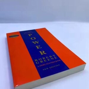 May include: A paperback book with a bright orange cover and a central vertical blue stripe. The title POWER by Robert Greene is printed in gold lettering on the blue stripe, with the text A Joost Elffers Book and New Edition below it.