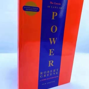 May include: A book titled The Concise 48 Laws of Power by Robert Greene and Joost Elffers. The cover features a vertical blue stripe centered on an orange background with gold text. A circular gold International Bestseller badge is at the top left.