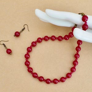 May include: A jewelry set consisting of a necklace and two matching drop earrings. The necklace features a single strand of round, glossy, deep red beads. Each earring consists of a single matching red bead suspended from a bronze-toned hook.