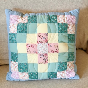 Puede incluir: Un cojín cuadrado con un diseño de patchwork tipo colcha y un patrón de cruz central. Los cuadrados de tela alternan entre amarillo pálido liso, verde claro liso y estampados florales en tonos rosa, verde y blanco, bordeados por azul claro.