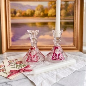 Könnte beinhalten: Zwei Kerzenhalter aus klarem Glas mit facettiertem Fuß und einem leuchtend magentafarbenen Blumenmuster. Ein Halter enthält eine lange weiße Stabkerze, während eine zweite weiße Stabkerze waagerecht auf einem weißen Tuch neben zwei Blumenkarten liegt.