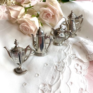 Könnte beinhalten: Ein Set aus vier silberfarbenen Metall-Serviettenringen, die jeweils ein Miniatur-Teekannen- oder Kaffeekannendesign mit detaillierten Prägungen aufweisen, angeordnet auf einem weißen Stoff mit mehreren blassrosa Rosenblüten.