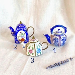 May include: Three miniature ceramic teapots labeled 2, 3, and 4. Teapot 2 is cobalt blue with a floral oval. Teapot 3 is white with pink and blue flowers and a gold handle. Teapot 4 is white with blue floral patterns and a blue handle.